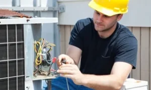 electrician wearing a yellow hard hat repairing the electrical wiring on an outdoors HVAC system